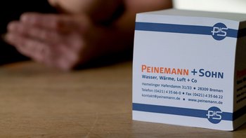 Eine weiße Box mit dem Logo von Peinemann + Sohn steht auf einem Holztisch, während eine Hand im Hintergrund sichtbar ist.