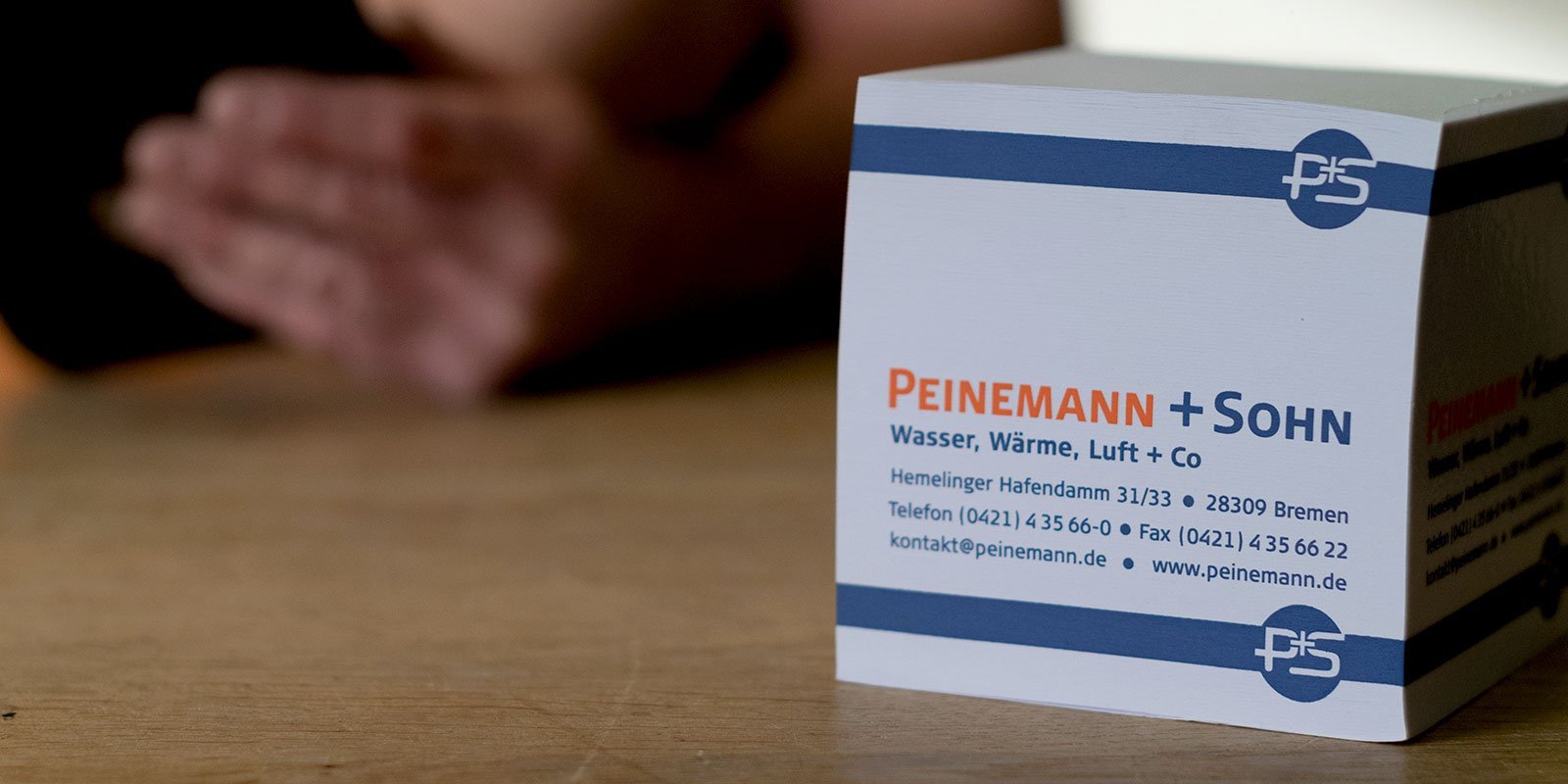 Eine weiße Box mit dem Logo von Peinemann + Sohn steht auf einem Holztisch, während eine Hand im Hintergrund sichtbar ist.